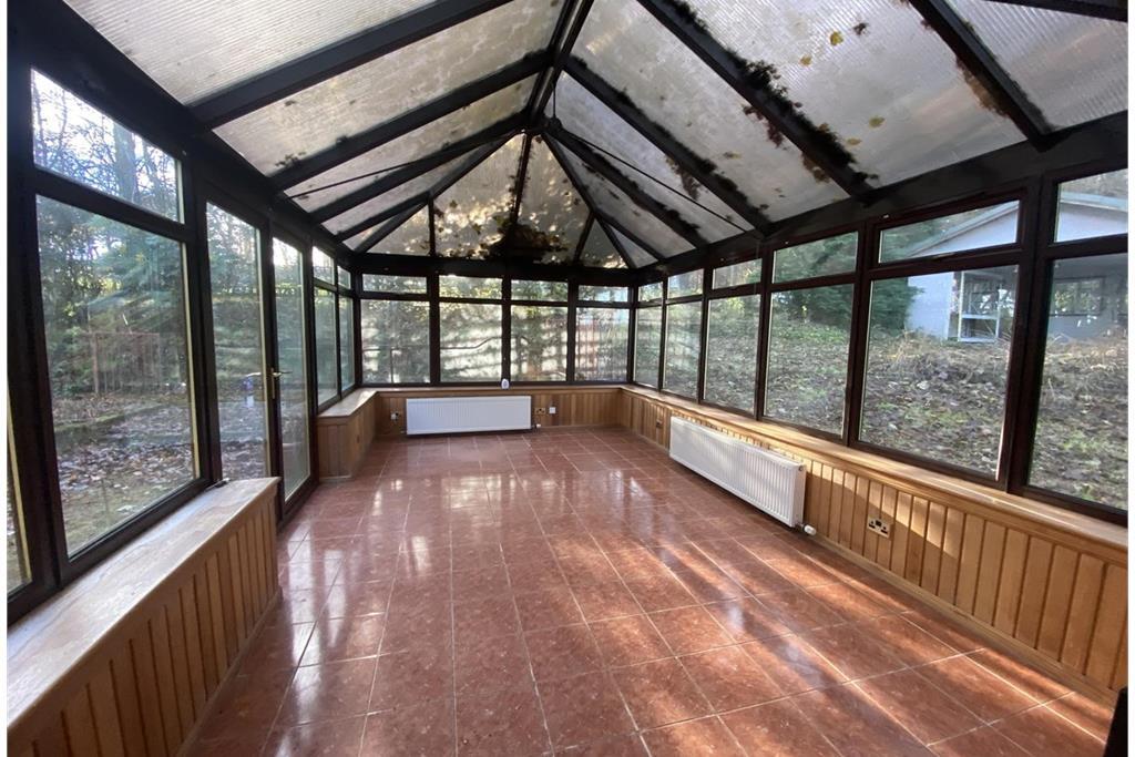 A large conservatory with black frames around the windows and red-brown floors. The garden outside is overgrown.